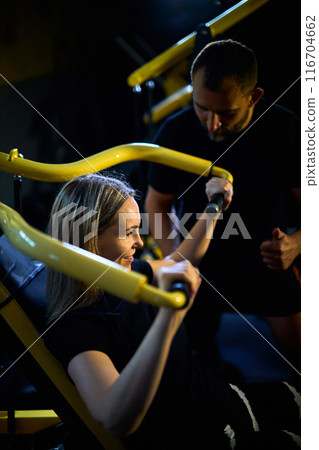 Personal coach assisting sportswoman in using machine 116704662