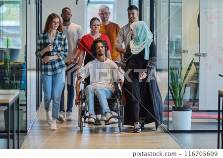 A diverse group of young business people walking a corridor in the glass-enclosed office of a modern startup, including a person in a wheelchair and a woman wearing a hijab, showing a dynamic mix of 116705609