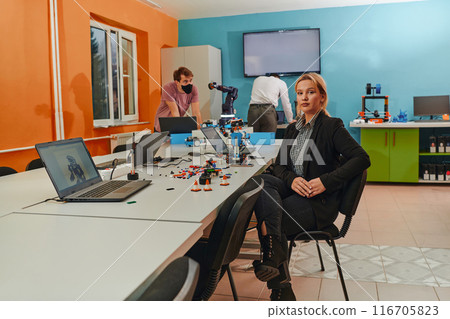 A woman sitting in a laboratory and solving problems and analyzing the robot's verification. In the background, colleagues are talking at an online meeting 116705823