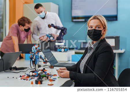 A woman wearing a protective mask standing in a laboratory while her colleagues test a new robotic invention in the background. A woman wearing a protective mask standing in a laboratory while her colleagues test a new robotic invention in the background. 116705854