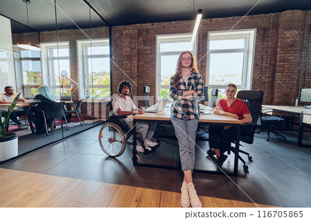 A young business group, including an African American businessman in a wheelchair, collaborates within a modern glass office, actively engaged around a computer and laptop, collectively solving 116705865