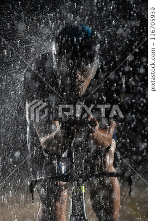 A triathlete braving the rain as he cycles through the night, preparing himself for the upcoming marathon. The blurred raindrops in the foreground and the dark, moody atmosphere in the background add A triathlete braving the rain as he cycles through the night, preparing himself for the upcoming marathon. The blurred raindrops in the foreground and the dark, moody atmosphere in the background add 116705939
