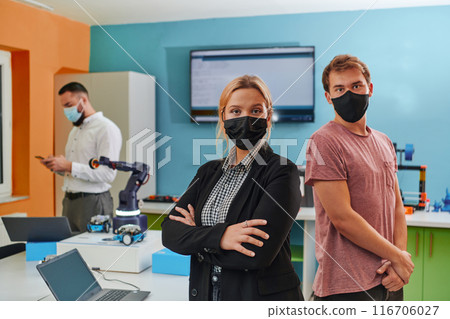 A woman wearing a protective mask standing in a laboratory while her colleagues test a new robotic invention in the background. A woman wearing a protective mask standing in a laboratory while her colleagues test a new robotic invention in the background. 116706027