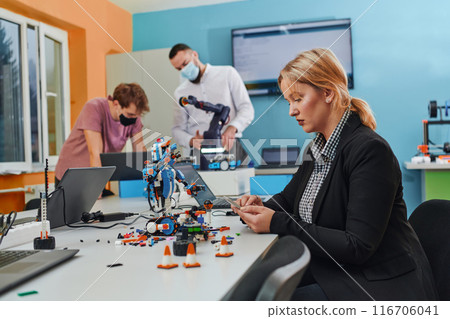 A woman sitting in a laboratory and solving problems and analyzing the robot's verification. In the background, colleagues are talking at an online meeting 116706041
