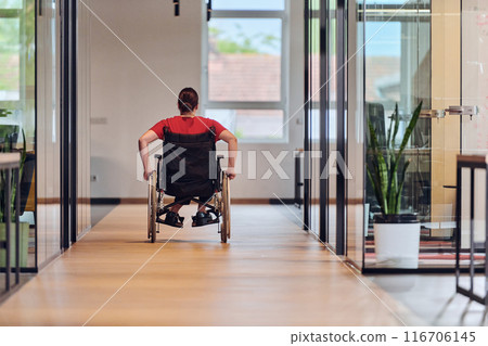A modern young businesswoman in a wheelchair is surrounded by an inclusive workspace with glass-walled offices, embodying determination and innovation in the business world 116706145