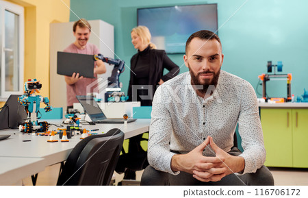 A man sitting in a robotics laboratory while his colleagues in the background test new, cutting edge robotic inventions. 116706172