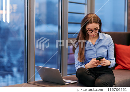 A businesswoman utilizes her laptop while seated by the window of a large corporate building, offering a picturesque view of the city skyline as her backdrop. A businesswoman utilizes her laptop while seated by the window of a large corporate building, offering a picturesque view of the city skyline as her backdrop. 116706261