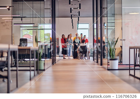 A diverse group of young business people congregates within a modern startup's glass-enclosed office, featuring inclusivity with a person in a wheelchair, an African American young man , and a hijab 116706326
