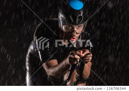 A triathlete braving the rain as he cycles through the night, preparing himself for the upcoming marathon. The blurred raindrops in the foreground and the dark, moody atmosphere in the background add 116706344
