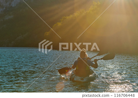 A young couple enjoying an idyllic kayak ride in the middle of a beautiful river surrounded by forest greenery in sunset time  116706448