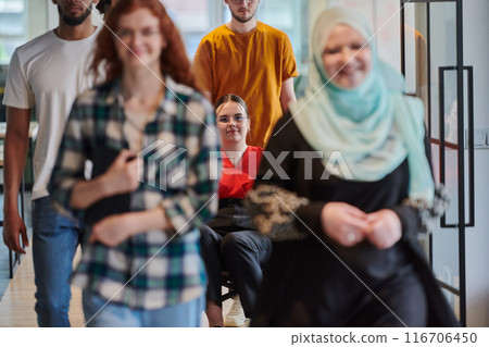 A diverse group of business people walking a corridor in the glass-enclosed office of a modern startup, including a person in a wheelchair and a woman wearing a hijab, showing a dynamic mix of 116706450