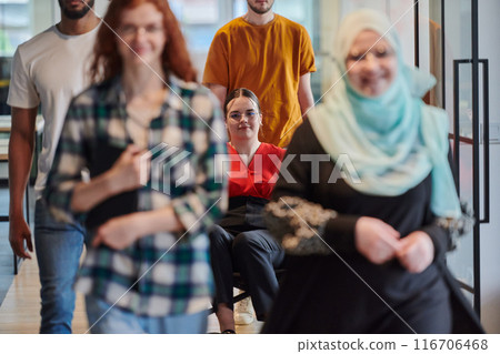 A diverse group of business people walking a corridor in the glass-enclosed office of a modern startup, including a person in a wheelchair and a woman wearing a hijab, showing a dynamic mix of 116706468