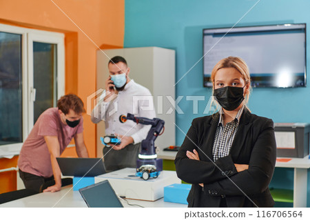 A woman wearing a protective mask standing in a laboratory while her colleagues test a new robotic invention in the background. A woman wearing a protective mask standing in a laboratory while her colleagues test a new robotic invention in the background. 116706564