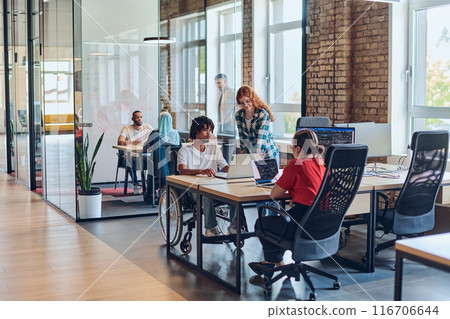 A young business group, including an African American businessman in a wheelchair, collaborates within a modern glass office, actively engaged around a computer and laptop, collectively solving A young business group, including an African American businessman in a wheelchair, collaborates within a modern glass office, actively engaged around a computer and laptop, collectively solving 116706644