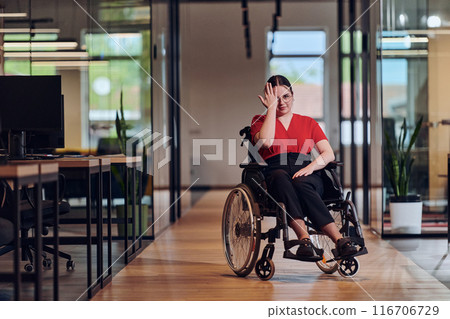 A modern young businesswoman in a wheelchair is surrounded by an inclusive workspace with glass-walled offices, embodying determination and innovation in the business world 116706729