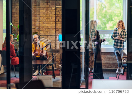A gathering of young business professionals, some seated in a glass-walled office, engage in a lively conversation and record an online podcast, embodying modern collaboration and dynamic interaction 116706733