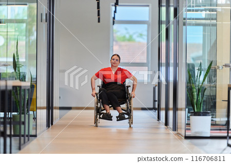 A modern young businesswoman in a wheelchair is surrounded by an inclusive workspace with glass-walled offices, embodying determination and innovation in the business world 116706811