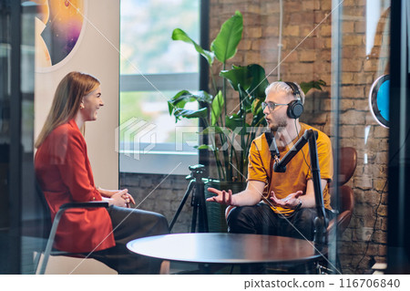 A gathering of young business professionals, some seated in a glass-walled office, engage in a lively conversation and record an online podcast, embodying modern collaboration and dynamic interaction 116706840
