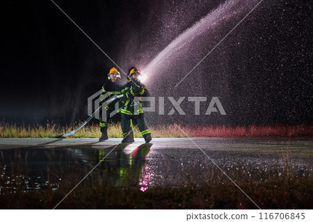 Firefighters using a water hose to eliminate a fire hazard. Team of female and male firemen in dangerous rescue mission.  116706845