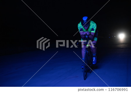 A triathlete rides his bike in the darkness of night, pushing himself to prepare for a marathon. The contrast between the darkness and the light of his bike creates a sense of drama and highlights the A triathlete rides his bike in the darkness of night, pushing himself to prepare for a marathon. The contrast between the darkness and the light of his bike creates a sense of drama and highlights the 116706991