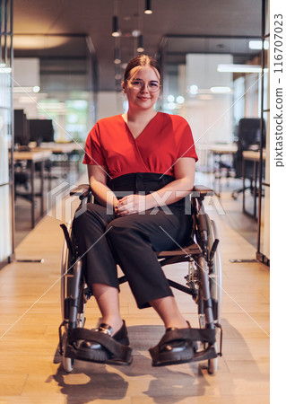 A modern young businesswoman in a wheelchair is surrounded by an inclusive workspace with glass-walled offices, embodying determination and innovation in the business world 116707023