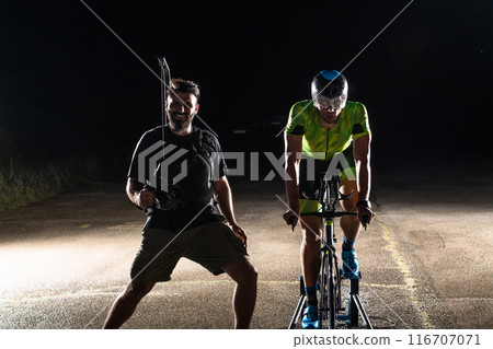 Behind the scenes. Professional videographer on a night shoot with a triathlete riding a bicycle for video recording purposes 116707071