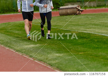 An inspiring and active elderly couple showcase their dedication to fitness as they running together on a lush green field, captured in a close-up shot of their legs in motion. 116707158