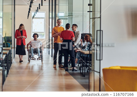 A group of young business people in a modern glass-walled office captures the essence of diversity and collaboration, while two colleagues, including an African American businessman in a wheelchair 116707166