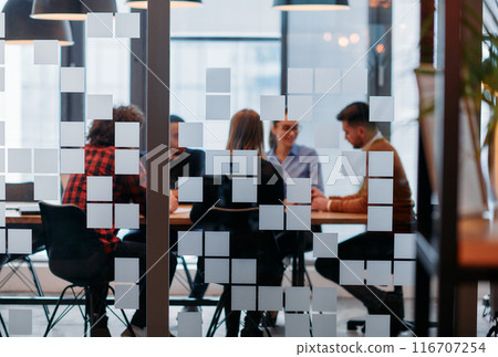 In a sleek glass office space, a team of business professionals engage in lively discussions and exchange strategic ideas, showcasing collaboration and innovation at the heart of modern corporate 116707254