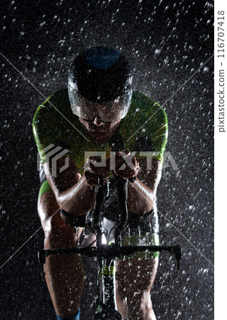 A triathlete braving the rain as he cycles through the night, preparing himself for the upcoming marathon. The blurred raindrops in the foreground and the dark, moody atmosphere in the background add A triathlete braving the rain as he cycles through the night, preparing himself for the upcoming marathon. The blurred raindrops in the foreground and the dark, moody atmosphere in the background add 116707418