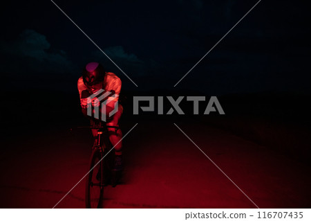 A triathlete rides his bike in the darkness of night, pushing himself to prepare for a marathon. The contrast between the darkness and the light of his bike creates a sense of drama and highlights the A triathlete rides his bike in the darkness of night, pushing himself to prepare for a marathon. The contrast between the darkness and the light of his bike creates a sense of drama and highlights the 116707435