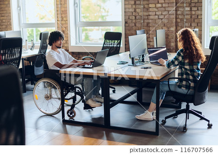 Business colleagues work within a modern startup coworking office, with one of them, an African America in a wheelchair, actively engaging with computers and laptops to collaboratively address various Business colleagues work within a modern startup coworking office, with one of them, an African America in a wheelchair, actively engaging with computers and laptops to collaboratively address various 116707566