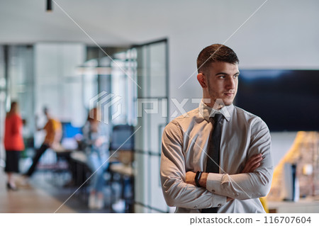 A young business leader stands with crossed arms in a modern office hallway, radiating confidence and a sense of purpose, embodying a dynamic and inspirational presence. 116707604