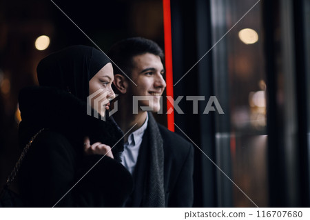 Happy multicultural business couple walking together outdoors in an urban city street at night near a jewelry shopping store window. Happy multicultural business couple walking together outdoors in an urban city street at night near a jewelry shopping store window. 116707680