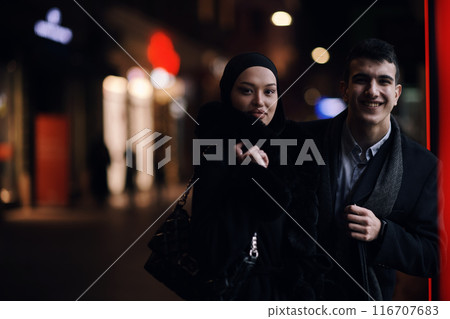 Happy multicultural business couple walking together outdoors in an urban city street at night near a jewelry shopping store window. Happy multicultural business couple walking together outdoors in an urban city street at night near a jewelry shopping store window. 116707683