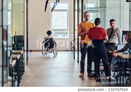 A diverse group of colleagues engages in a discussion about business challenges within a modern coworking startup center, while in the background, their wheelchair-bound colleague symbolizes A diverse group of colleagues engages in a discussion about business challenges within a modern coworking startup center, while in the background, their wheelchair-bound colleague symbolizes 116707684