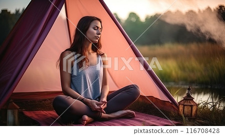 A young woman meditates early in the morning in the summer forest near her tent. 116708428