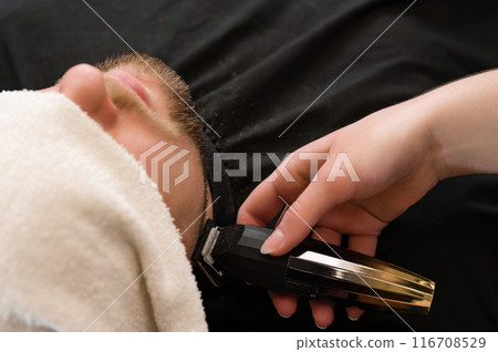 The clients head is covered with a towel during a beard trim. Shortening the length of the beard from the sides by the master for the client. The clients head is covered with a towel during a beard trim. Shortening the length of the beard from the sides by the master for the client. 116708529