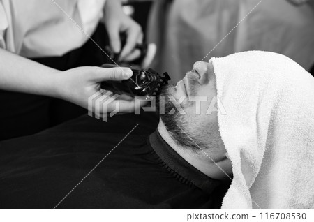 The clients head is covered with a towel during a beard trim. Shortening the length of the beard from the sides by the master for the client. The clients head is covered with a towel during a beard trim. Shortening the length of the beard from the sides by the master for the client. 116708530