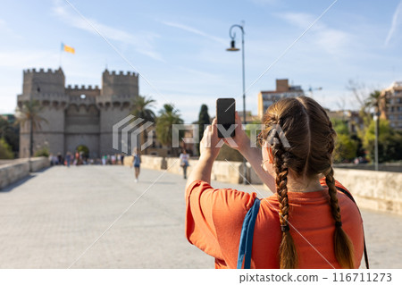 Redhead female traveler in casual dress taking picture of Torres de Serranos tower with smartphone, travel in Europe. Attractive female tourist is exploring new city. Hight quality photo, rear view Redhead female traveler in casual dress taking picture of Torres de Serranos tower with smartphone, travel in Europe. Attractive female tourist is exploring new city. Hight quality photo, rear view 116711273