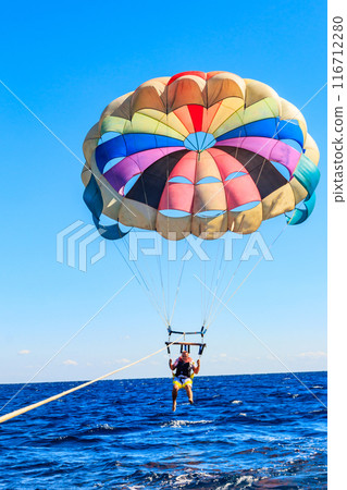 Parasailing water amusement - flying on a parachute behind a boat on a summer holiday by the sea. Summer vacation concept Parasailing water amusement - flying on a parachute behind a boat on a summer holiday by the sea. Summer vacation concept 116712280