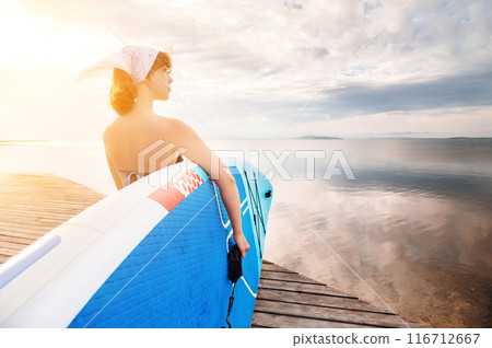 Young beautiful woman in a bikini holds a SUP board on the sea beach. The girl carries a board against the background of the sun and smiles 116712667