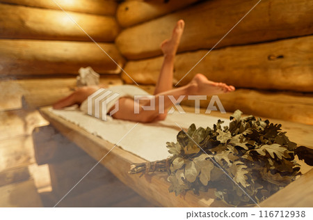Young woman relaxing and sweating in hot sauna wrapped in towel. Girl In Sauna. Interior of Finnish sauna, classic wooden steam room with hot steam. Russian bathroom. Relax in hot Bathhouse with steam 116712938