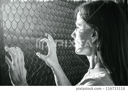 A woman stands behind a chainlink fence, in black and white, showing contemplation and separation A woman stands behind a chainlink fence, in black and white, showing contemplation and separation 116715118