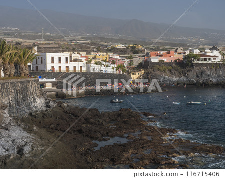 Views of the picturesque Alcala village with traditional architecture houses, small tranquil cove with beach and pier crowded with sunbathing people. Tenerife, Canary Islands, Spain. Views of the picturesque Alcala village with traditional architecture houses, small tranquil cove with beach and pier crowded with sunbathing people. Tenerife, Canary Islands, Spain. 116715406