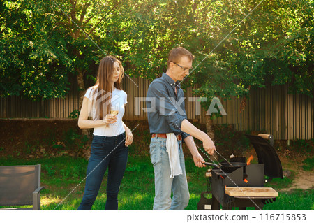Middle-aged man grilling a steak on a grill and his daughter watching on him standing at backyard Middle-aged man grilling a steak on a grill and his daughter watching on him standing at backyard 116715853