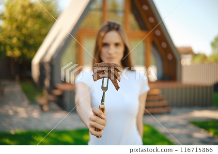 Defocused woman stretching forward a barbecue fork with slices of steak in her hand 116715866