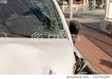 Close-up of a white car with significant front damage and shattered windshield, highlighting the aftermath of a road collision. Useful for insurance, safety campaigns, or automotive repair content. Close-up of a white car with significant front damage and shattered windshield, highlighting the aftermath of a road collision. Useful for insurance, safety campaigns, or automotive repair content. 116716169