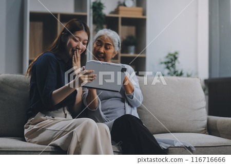 Happy grownup daughter showing content on tablet to mature mother. Two family generations women with digital computer gadget resting on couch together, watching videos, making video call Happy grownup daughter showing content on tablet to mature mother. Two family generations women with digital computer gadget resting on couch together, watching videos, making video call 116716666
