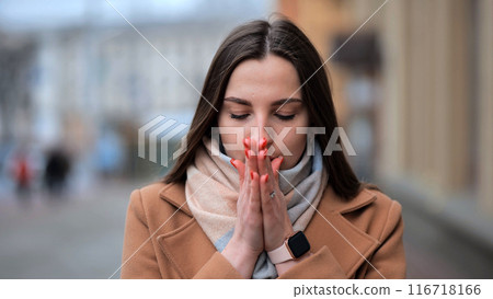 A girl warms her palms with her mouth in the cold time in the city. 116718166